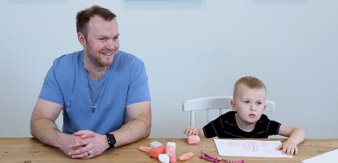 Evan and son Malachi sit at table with coloring paper while smiling.